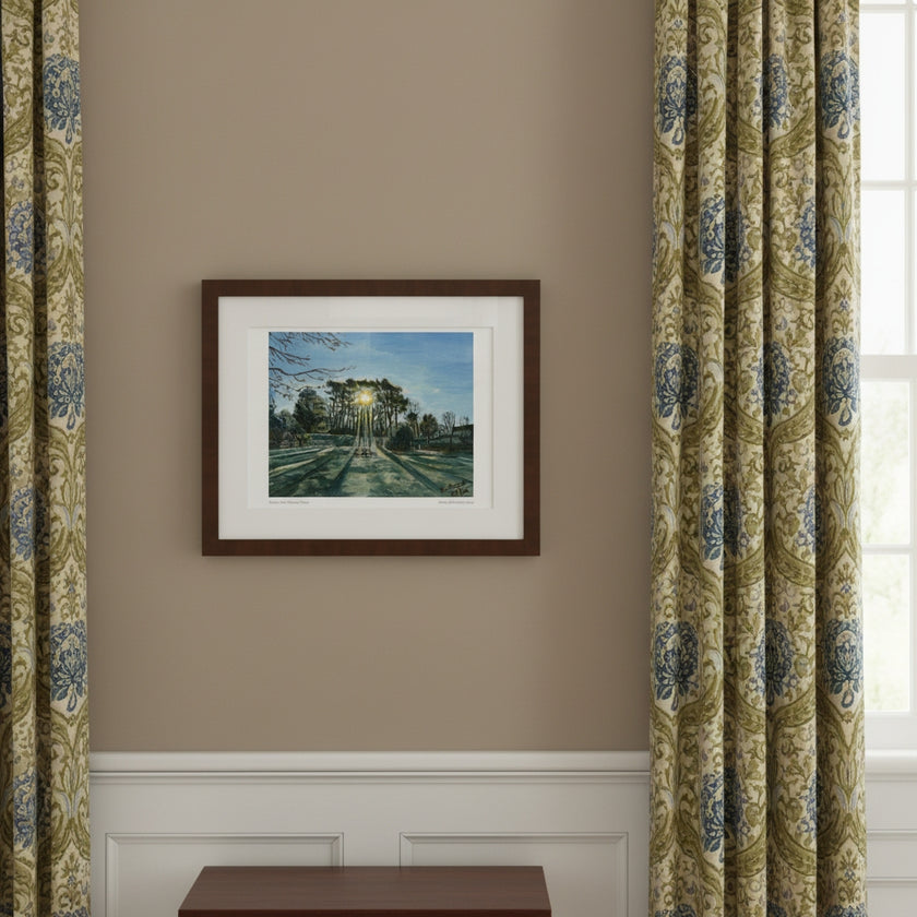 A framed art print depicting a sunlit forest path hangs on a beige wall between two light blue curtains. A small round wooden table below holds a mug, papers, and stacked books.