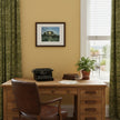 A framed art print depicting a forest path with dappled sunlight and shadows, hung on a dark blue wall in a dining room setting with a wooden table, chairs, and built-in shelves with copper pots and books.
