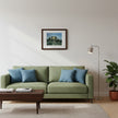 A framed art print depicting a forest path hangs on a mustard yellow wall above a brown leather sofa with green and blue throw pillows. A wooden coffee table with books and a mug sits in the foreground.