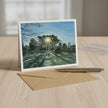A landscape art print card displayed on a light wood table, standing upright next to a brown kraft envelope and a silver pen. The card features a painting of a park scene with a bright sun shining through trees.