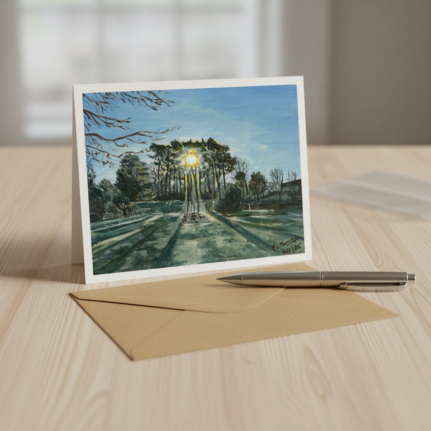 A landscape art print card displayed on a light wood table, standing upright next to a brown kraft envelope and a silver pen. The card features a painting of a park scene with a bright sun shining through trees.