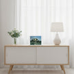 A greetings card showing a painting of sun coming through trees on a white sideboard with a vase of white flowers nd a white lamp