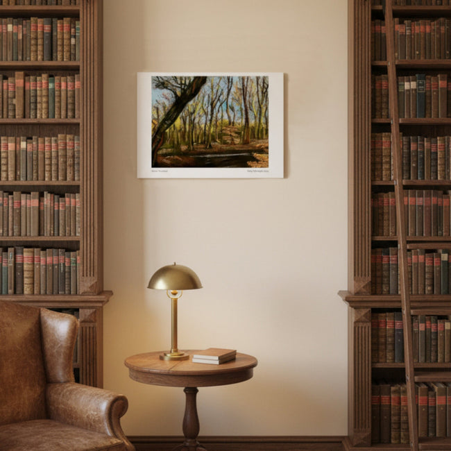 A framed landscape art print depicting a forest path with autumn trees, displayed on a beige wall in a cosy library setting with bookshelves, a leather armchair, and a brass table lamp.