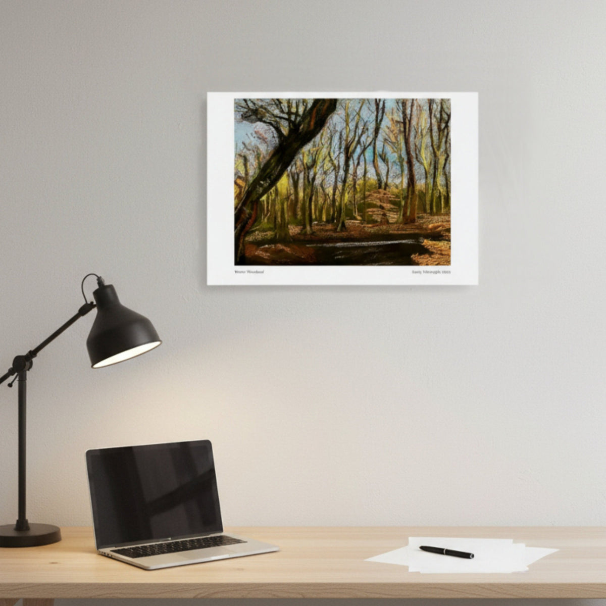A framed art print depicting a forest scene with autumn trees, displayed on a light grey wall above a wooden desk with a laptop, black desk lamp, and papers.