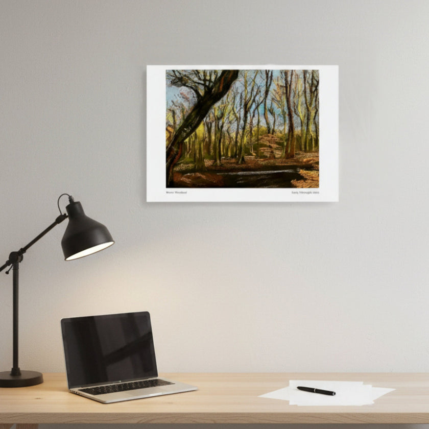 A framed art print depicting a forest scene with autumn trees, displayed on a light grey wall above a wooden desk with a laptop, black desk lamp, and papers.