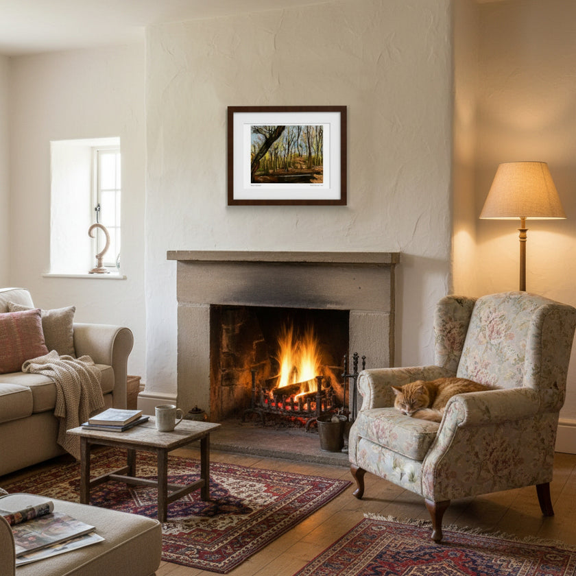 A framed landscape art print depicting a forest is displayed on a wall above a fireplace in a cosy, warmly lit living room with a cat resting in an armchair.