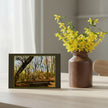 A greetings card depicting a forest scene with bare trees , styled on a light wood table next to a brown ceramic vase filled with yellow flowers.