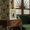 A greetings card of Ginger Rogers, styled on a wooden side table in a modern room with patterned wallpaper and a green armchair.